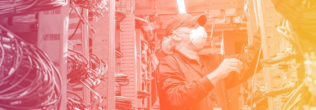 A masked technician attends to a wall of technology equipment, adjusting cables.